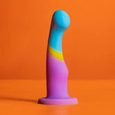 The Avant D14 G-Spot or Prostate Dildo – Heart of Gold by Avant, a colorful platinum cured silicone toy with a curved shape in blue, yellow, and purple, stands upright on an orange surface against an orange background.